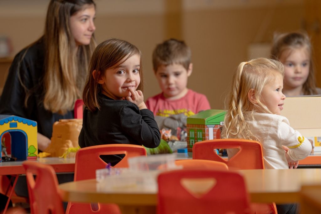 Children playing with toys in a classroom, with a teacher in the background