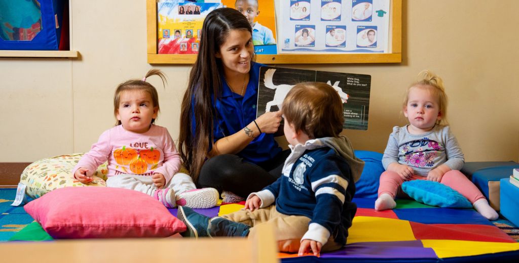 A caregiver in a blue shirt sits on a colorful mat reading a book to three toddlers in a bright childcare classroom