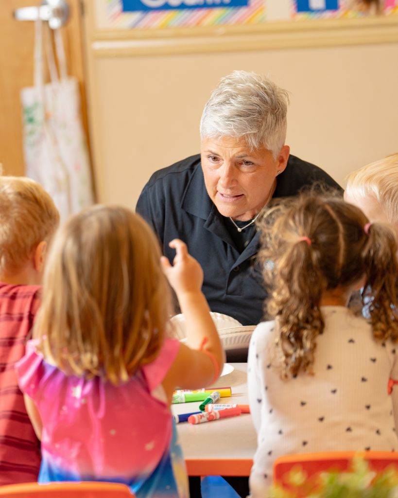 An experienced caregiver leans in attentively while engaging with young children seated at a table with crayons and art supplies
