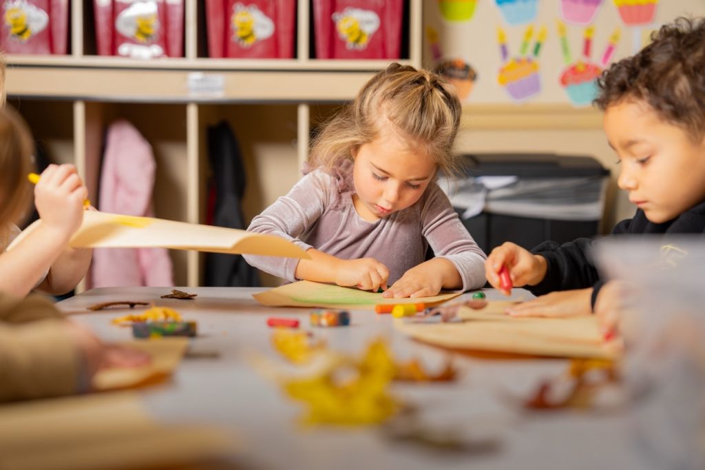 Children focused on an art and craft activity at a table in a colorful classroom with cubbies and birthday cake decorations in the background