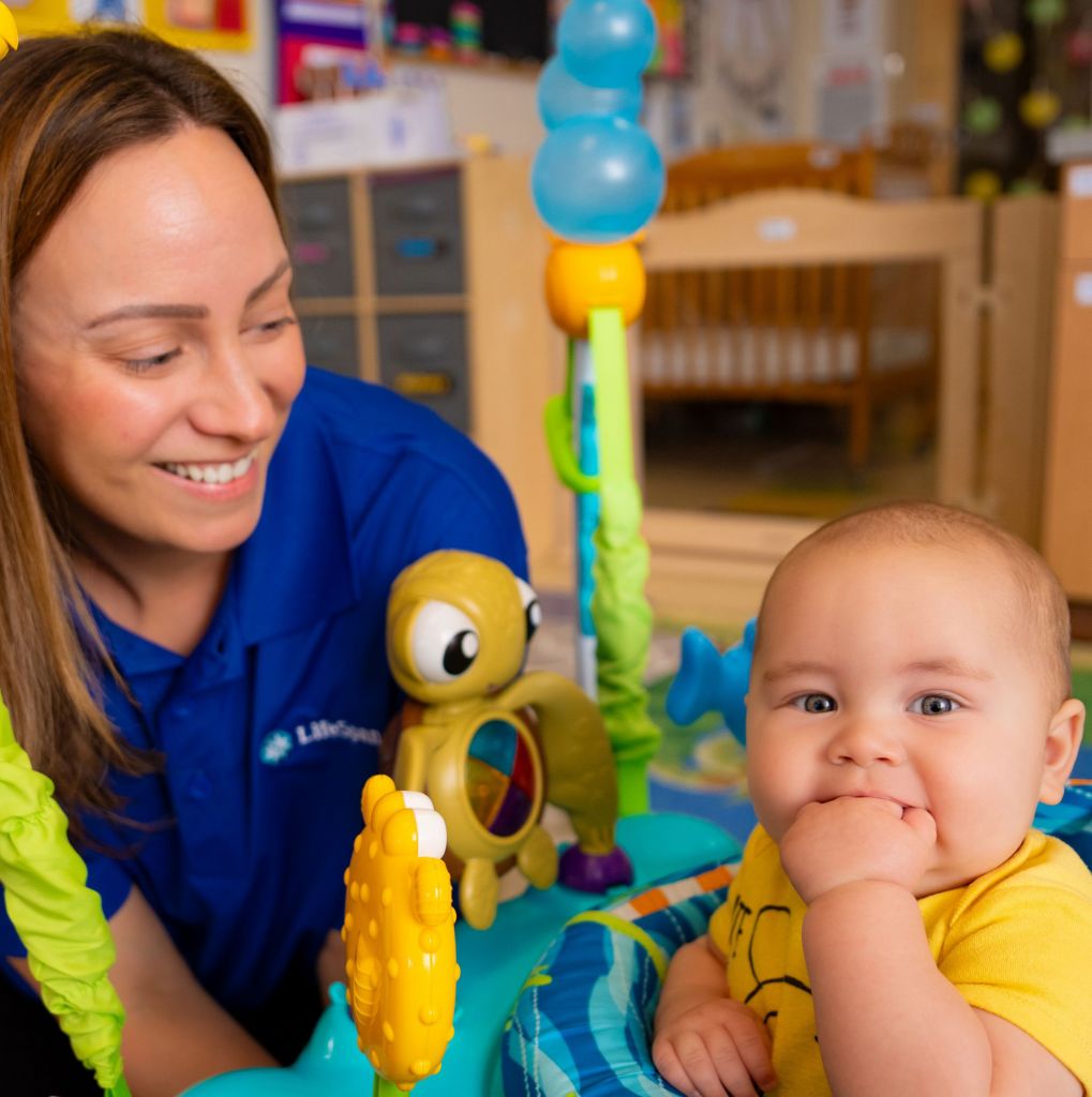 A smiling Lifespan caregiver in a blue shirt interacts with a baby sitting in a colorful toy activity center in a childcare room