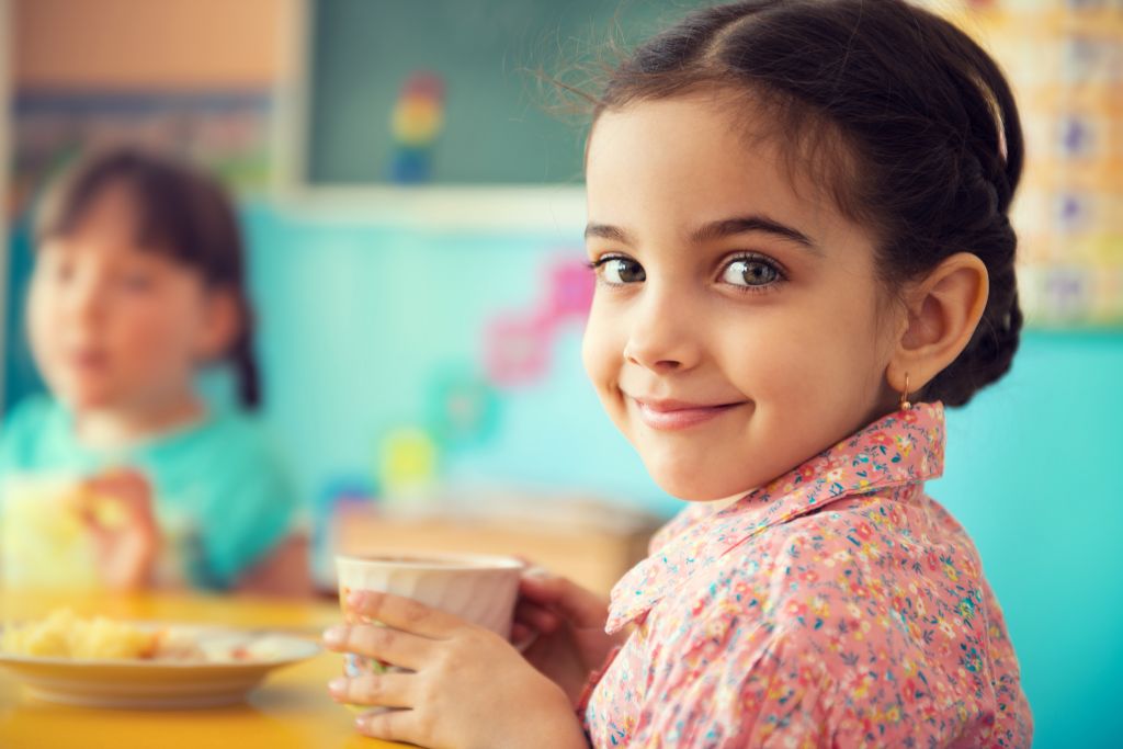 A young girl smiles at the camera while holding a cup, seated at a table in a colorful classroom with another child in the background