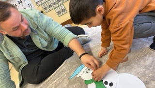 A caregiver and young child sit on the floor together engaged in a hands-on craft activity