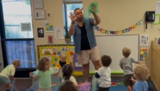 A teacher stands at the front of a colorful classroom while young children sit on the floor looking up attentively