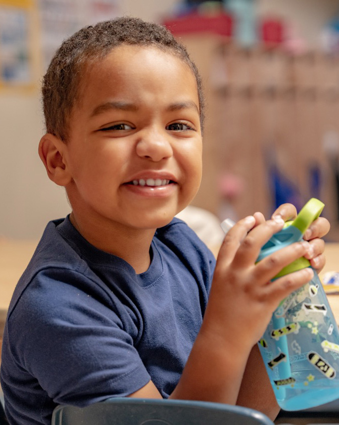 A smiling young boy holds a water bottle while seated at a table in a childcare classroom