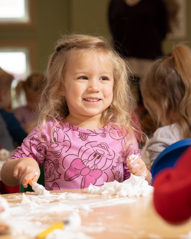 A smiling young girl in a pink shirt plays with foam or shaving cream at a table during sensory play in a childcare setting