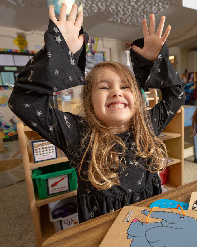 A happy young girl raises both hands high with a big smile while standing in a childcare classroom with bookshelves and learning materials behind her