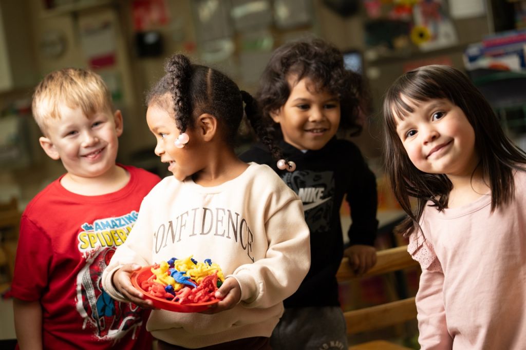 Four young children smile together in a childcare classroom, one holding a bowl of colorful toy figures