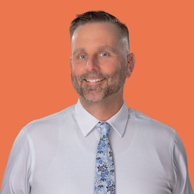 A Lifespan Childcare staff member with short gray-streaked hair and a beard wearing a white shirt and floral tie, smiling against an orange background